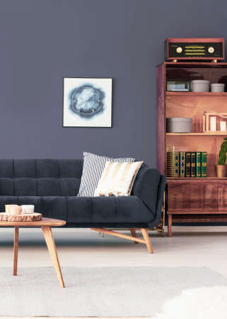 Wooden cabinet next to couch in dark apartment interior with poster and table. Real photo