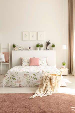 Blanket and pillows on bed with plants on headboard in pink and white bedroom interior. Real photo
