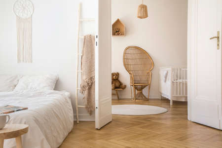 White apartment interior with herringbone parquet, double bed and open door to kid room with peacock chair, white crib and round rug on the floor White apartment interior with herringbone parquet, double bed and open door to kid room with peacock chair, white crib and round rug on the floor
