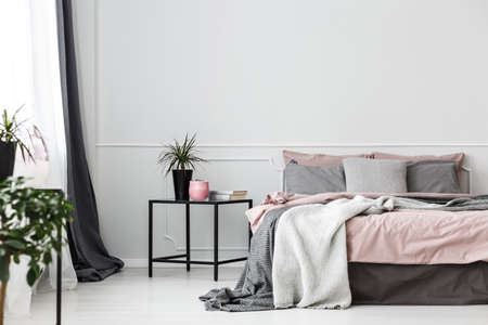 Black, box frame side table next to a cozy bed with gray blankets and pink sheets in an elegant bedroom interiorの写真素材