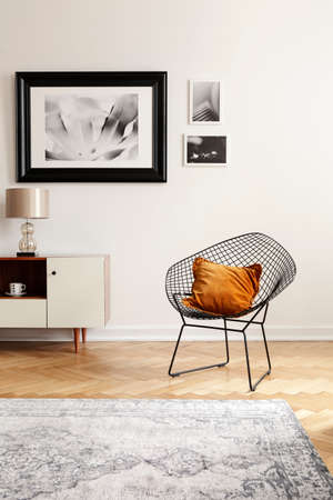 Orange pillow on a black, industrial net chair by a white wall with gallery of mock-up pictures in an elegant living room interior.の写真素材