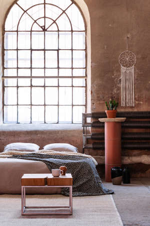Table in front of bed with cushions in bedroom interior with window and plant. Real photo