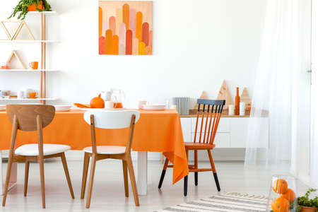 Real photo of kitchen table covered with orange tablecloth and white dishes. Artwork on the wall and...