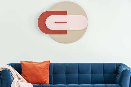 Modern poster on white wall above blue sofa with orange cushion and pink blanket in interior. Real...
