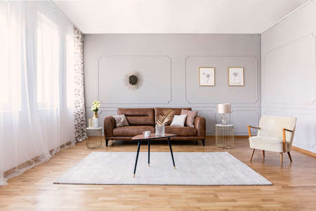 Brown leather sofa in elegant living room interior with stylish armchair, coffee table and drawings...