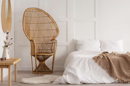 Flowers on wooden table next to rattan chair in white bedroom interior with blanket on bed. Real...
