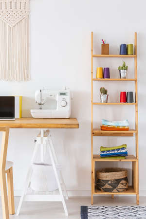 Real photo of a sewing machine on a desk next to a wooden shelf with colorful threadsの写真素材