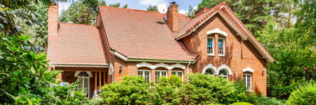 A red brick English style classic house with a steep roof and large windows surrounded by trees and green plants. Panorama.の写真素材