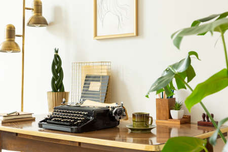 Typewriter, cup of coffee and plant on wooden desk in workspace interior with gold lamp. Real photo