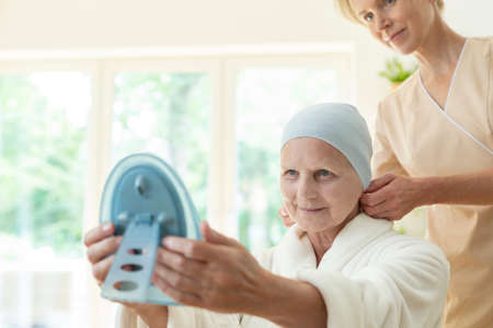 Happy sick elderly woman with headscarf and caregiver looking in the mirror