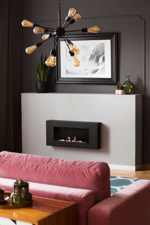 Lamp above pink settee in grey modern living room interior with poster above fireplace. Real photo