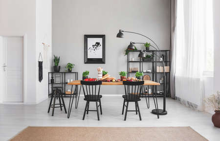 Black chairs at dining table with food in apartment interior with lamp and poster on grey wall. Real...