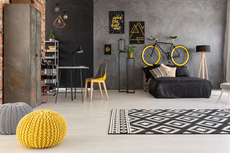 Yellow pouf and patterned carpet in grey teenager's room with bike above black bed. Real photoの写真素材