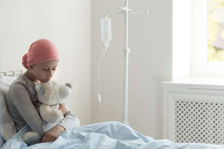 Sad ill teenager girl sitting alone in hospice bed, holding teddy bear, waiting for her doctorの写真素材
