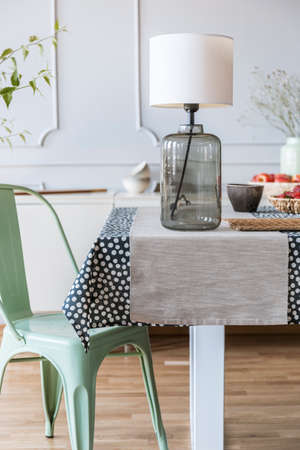 Close-up of a glass lamp on a table next to a green chair in a daily room interior. Real photo