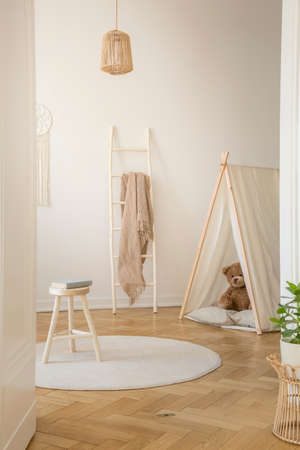 Vertical view of white scandinavian playroom with tent, teddy bear and wooden ladder with beige...