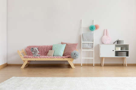 Comfortable sofa with pink blanket and colorful pillows next to white wooden ladder and shelf with round boxes, real photo with copy space on the white wallの写真素材