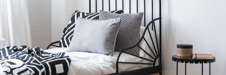 Close-up of bedside table with box and notebook placed next to black metal bed with grey pillows and...