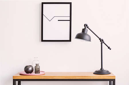 Closeup of table with lamp and plate with glass bottles, poster in black frame on white wall aboveの写真素材