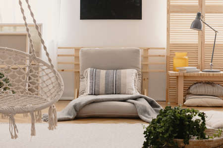 Pillow and blanket on grey futon in boho bedroom interior with lamp and hanging chair. Real photoの写真素材