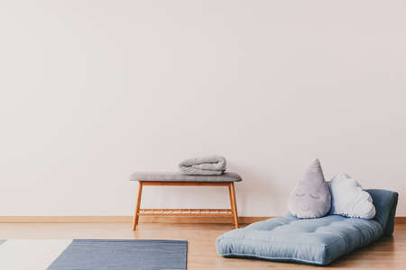 Blue futon with cute pillows on the floor of stylish baby room interior with copy space on the empty white wall and wooden and grey table with lampの写真素材