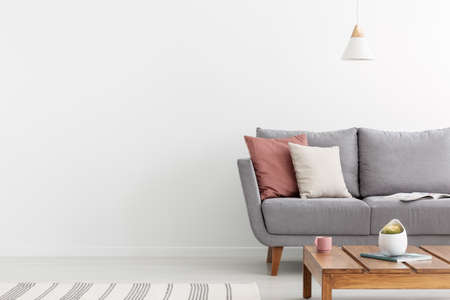 Pillows on grey settee and wooden table in white apartment interior with copy space on the wall. Real photoの写真素材