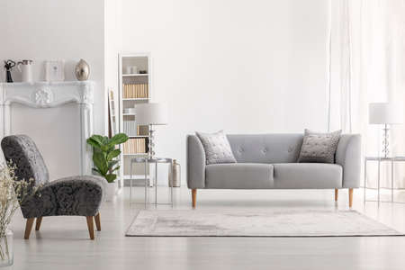 Carpet near armchair and grey settee in white living room interior with lamp on table. Real photoの写真素材