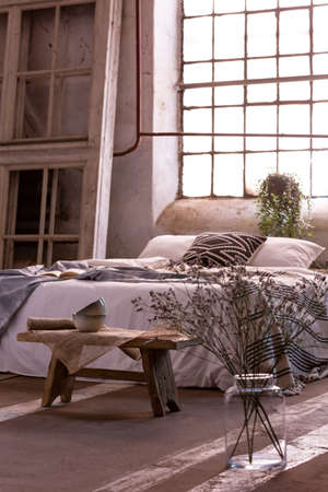 Flowers next to wooden bench in bright loft bedroom interior with window behind bed. Real photoの写真素材