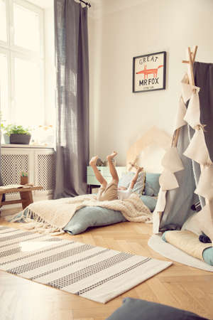 Kid with wooden aeroplane playing on blue bed in bright bedroom interior with tent and rug. Real photoの写真素材