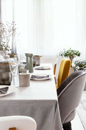 Real photo of table with dinnerware, candle and grey and yellow chair standing in bright dining room interior with window with curtainsの写真素材