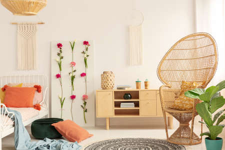 Wicker peacock chair with patterned pillow in cozy bedroom interior with macrame on the wall, flower...