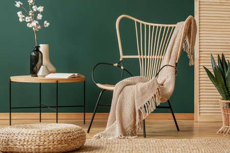 Pouf in front of table with flowers next to armchair with blanket in green loft interior. Real photo