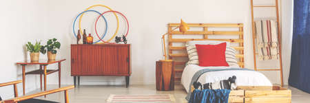 Bright teenager bedroom in real photo with retro cupboard with bottles, bed with wooden bedhead, and...