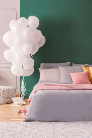 Dark green wall with copy space in colorful bedroom with bunch of white balloons next to double bed...