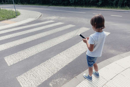 Boy with a smartphone standing at pedestrian crossing next to the road