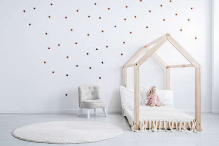 Pastel grey armchair next to wooden house shape bed with toy and blanket, copy space and golden...
