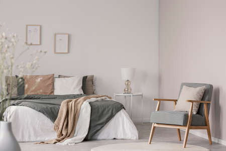 Wooden armchair next to bed with blanket and cushions in white bedroom interior. Real photoの写真素材