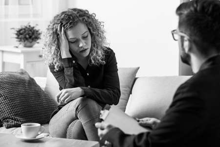 Black and white pretty blonde worried teenage girl sitting on the couch in psychotherapist office
