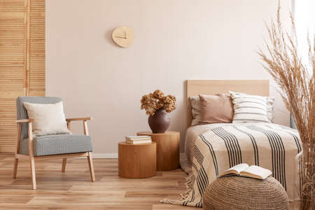 Stylish retro armchair with white pillow next to two round wooden tables with books and vase with flowers in beige bedroom interiorの写真素材