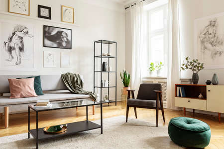 Pouf next to armchair in living room interior with table in front of sofa under posters. Real photo