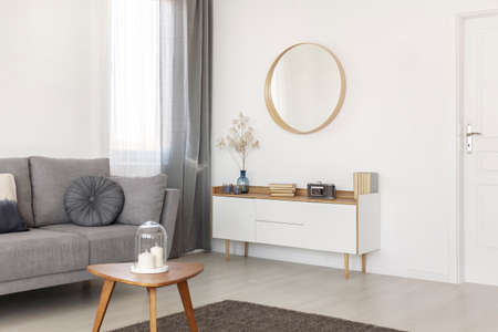 Stylish round mirror in wooden frame on white empty wall of elegant living room with cabinet, coffee table and sofaの写真素材