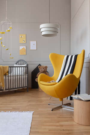 Striped black and white blanket on fancy yellow egg chair in cute baby room with posters on the wall and wooden cribの写真素材