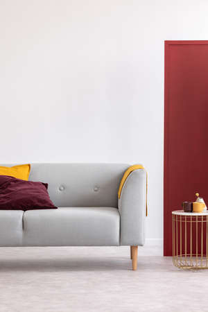 Gold table next to grey settee with pillows and blanket in white and red living room interior. Real photoの写真素材