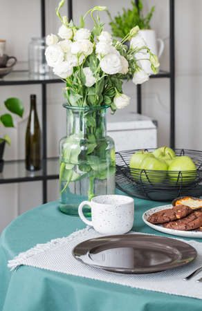 White roses in glass vase next to coffee mug and big plate on dining table