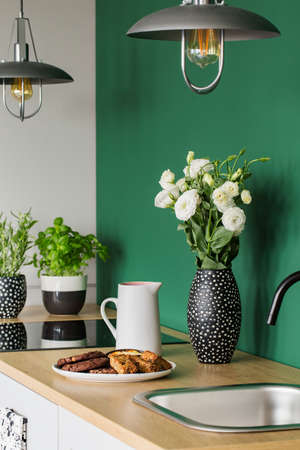 White roses in black and white vase next to coffee jug and big plate with cookies on wooden counter topの写真素材