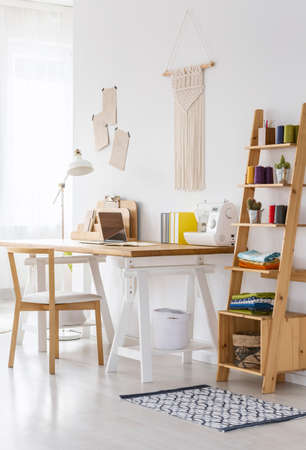 Wooden organizer next to desk with sewing machine in workspace interior with chair. Real photo