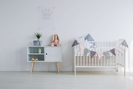 White wooden crib with pillows next to wooden cabinet with toy and green pant in grey, copy space on empty white wallの写真素材