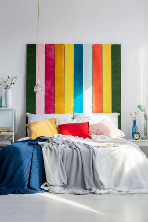 Rainbow bedhead behind white bed with colorful pillows and blankets in bright bedroom interior. Real photoの写真素材