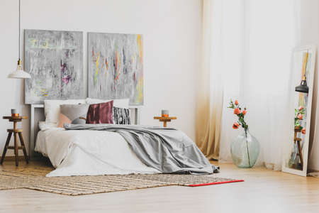 Natural rug on wooden floor of stylish bedroom interior with king size bed, wooden nightstands and grey fancy paintings on white wallの写真素材