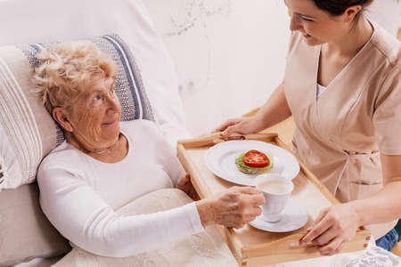 Helpful volunteer in beige uniform serving coffee to senior female patientの写真素材